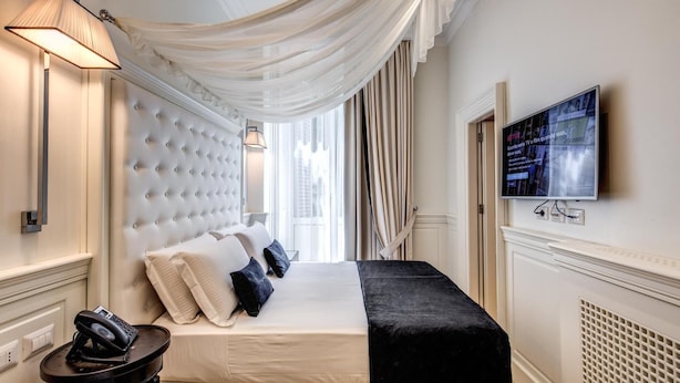 A hotel room with a tufted headboard, a bed with white and black bedding, a bedside telephone, a television on the wall, and sheer drapes. - Hotel dei Quiriti Suite (Rome)