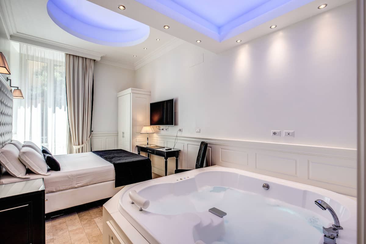 A modern bedroom with a large white bathtub, a bed with white linens, a desk with a lamp, and a TV on the wall.