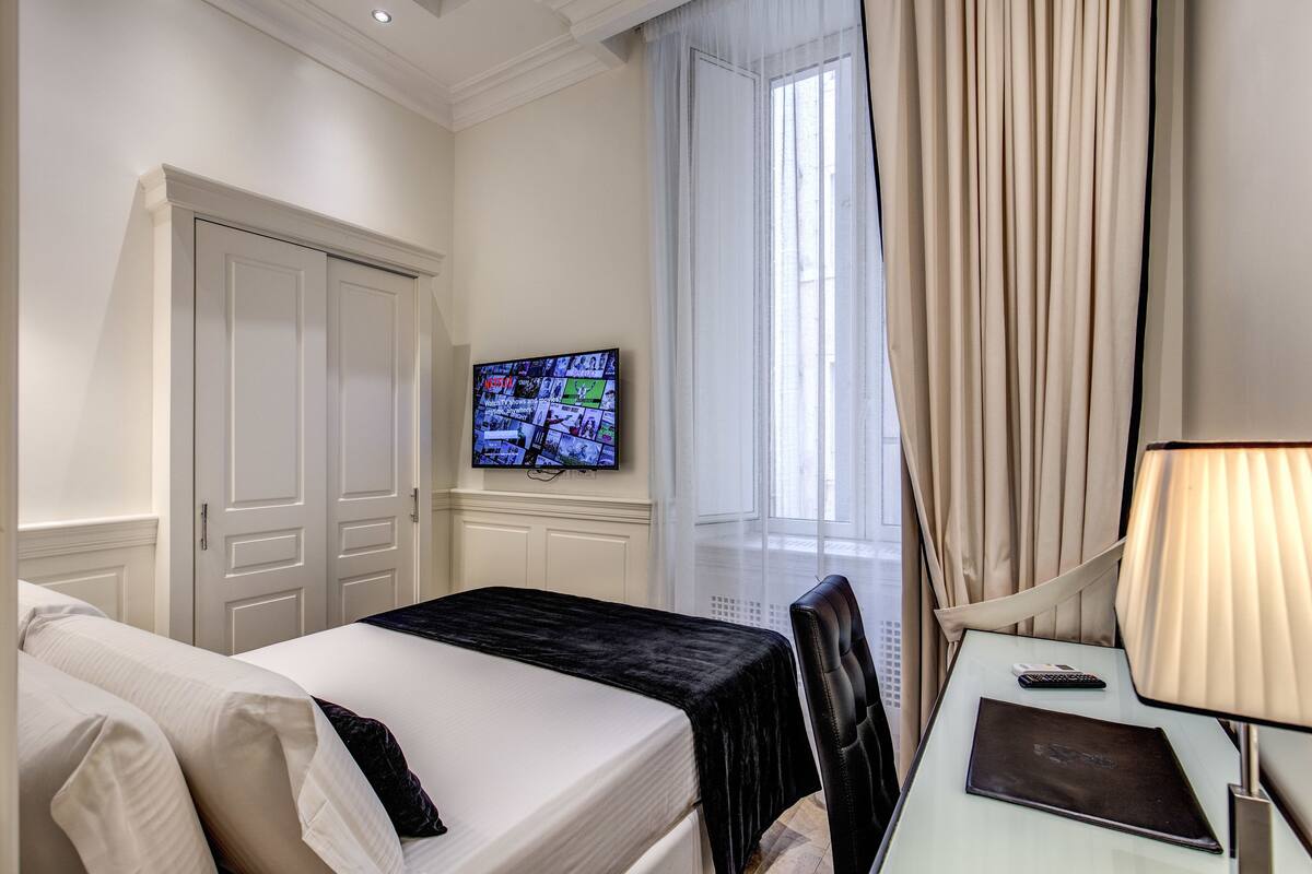 A hotel room with a bed, a desk with a lamp, a television on the wall, and a window with curtains.