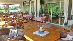 Un amplio espacio interior con varias mesas y sillas para comer, ventanas grandes que ofrecen vistas al exterior y una zona de estar central con una mesa redonda.