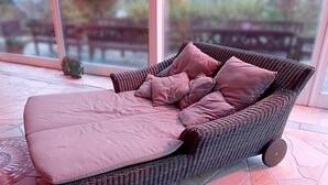 Un sofá de mimbre con un cojín rosa y ruedas, ubicado en un patio empedrado con vistas a mobiliario exterior.