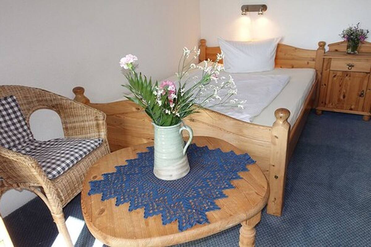 A wooden bed with a white mattress, a wicker chair with a checkered cushion, and a small round table with a blue doily.