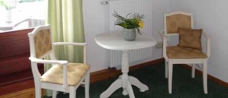 Una stanza con un tappeto verde, due sedie bianche, un piccolo tavolo bianco con un vaso, una televisione su un mobile in legno e una finestra con tende verdi.