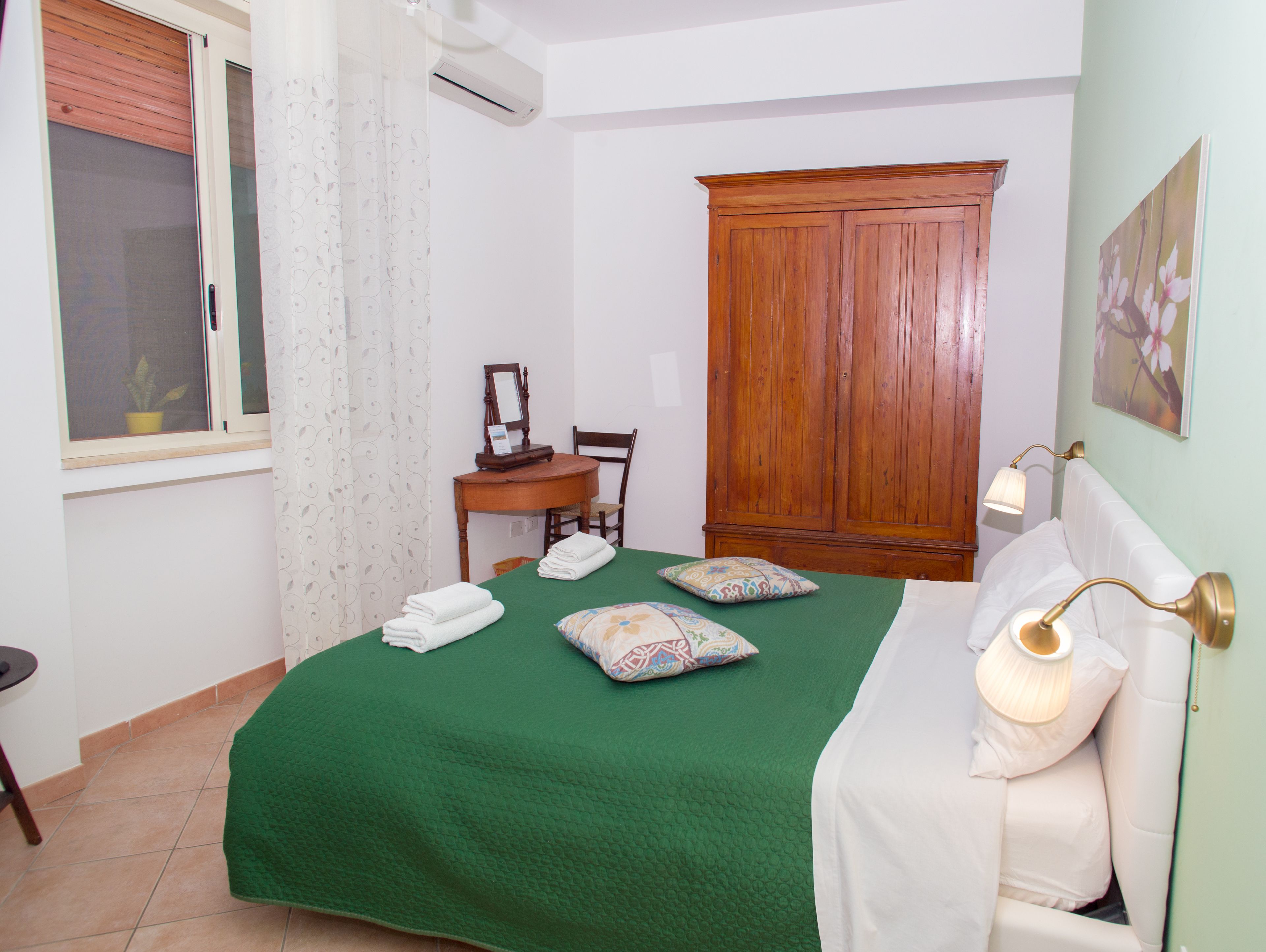 Un dormitorio ordenado con colcha verde, un armario de madera, un escritorio con silla y una lámpara de noche.