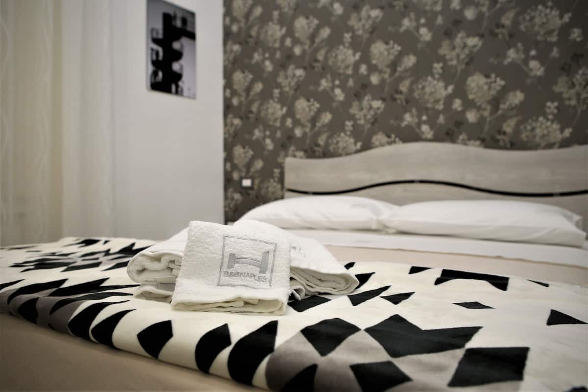 A neatly made bed with a patterned bedspread, a folded towel with a logo, and a floral wallpapered headboard.