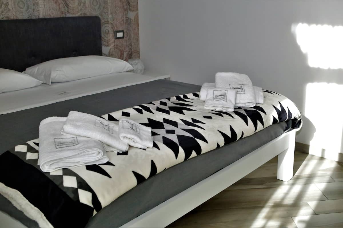 A neatly made bed with a patterned bedspread, a folded towel, and a pillow on a white bed frame in a room with a patterned wall and light-colored flooring.