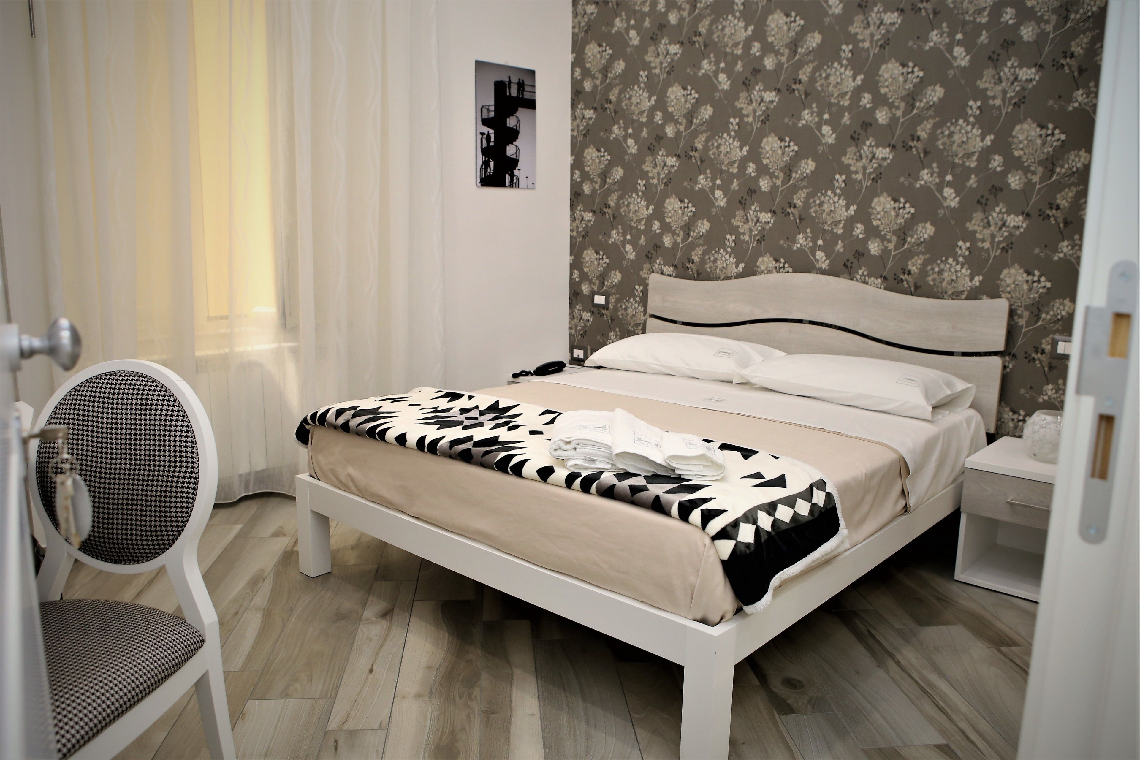 A neatly arranged bedroom with a bed, a chair, a nightstand, and a wall with a floral pattern.