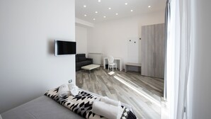 A modern hotel room with a sofa, a TV mounted on the wall, a small table, and a chair. - Hermes Rooms for tourists (Naples)