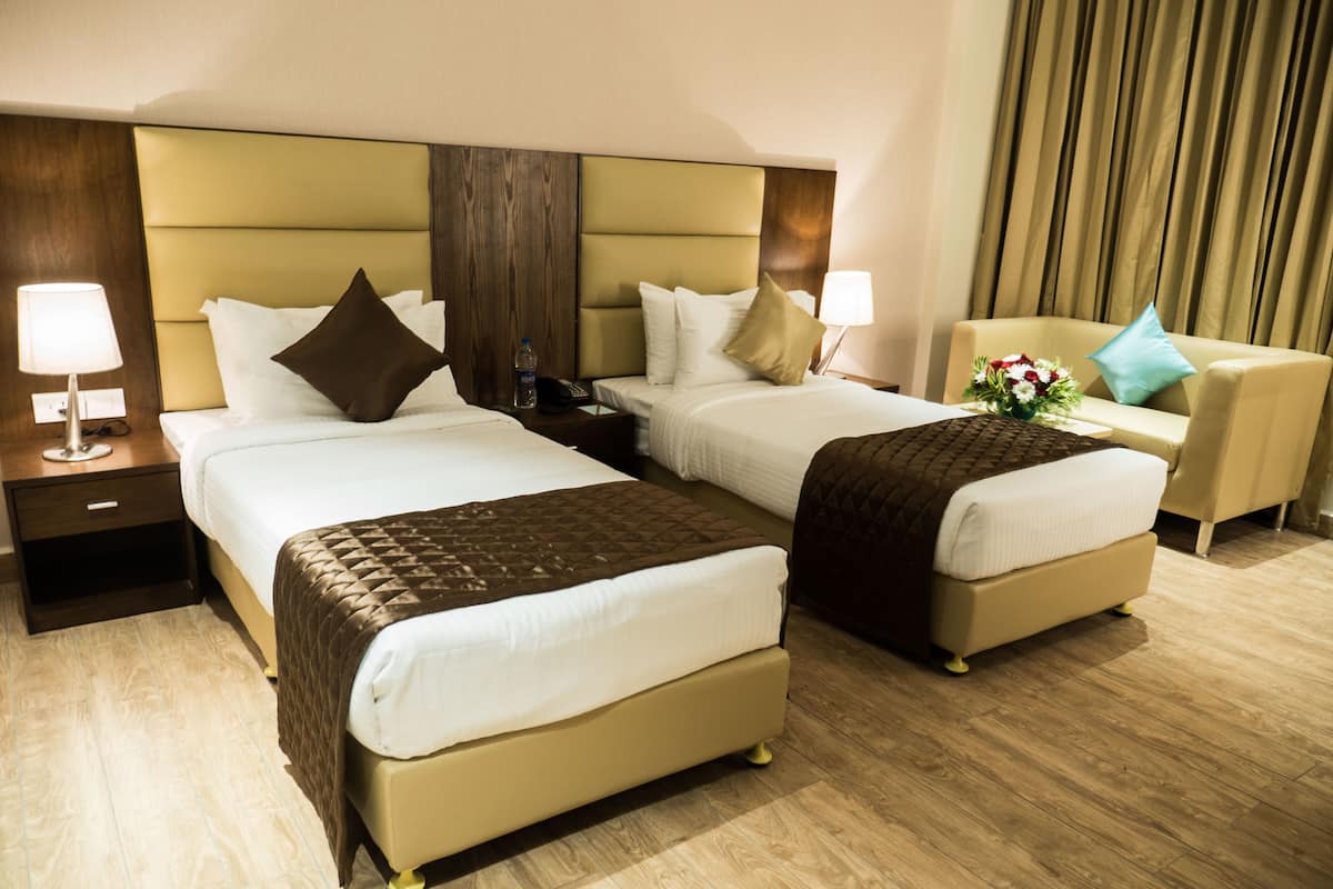 Habitación de hotel con dos camas, una mesita de noche con lámpara, una silla y un jarrón decorativo.