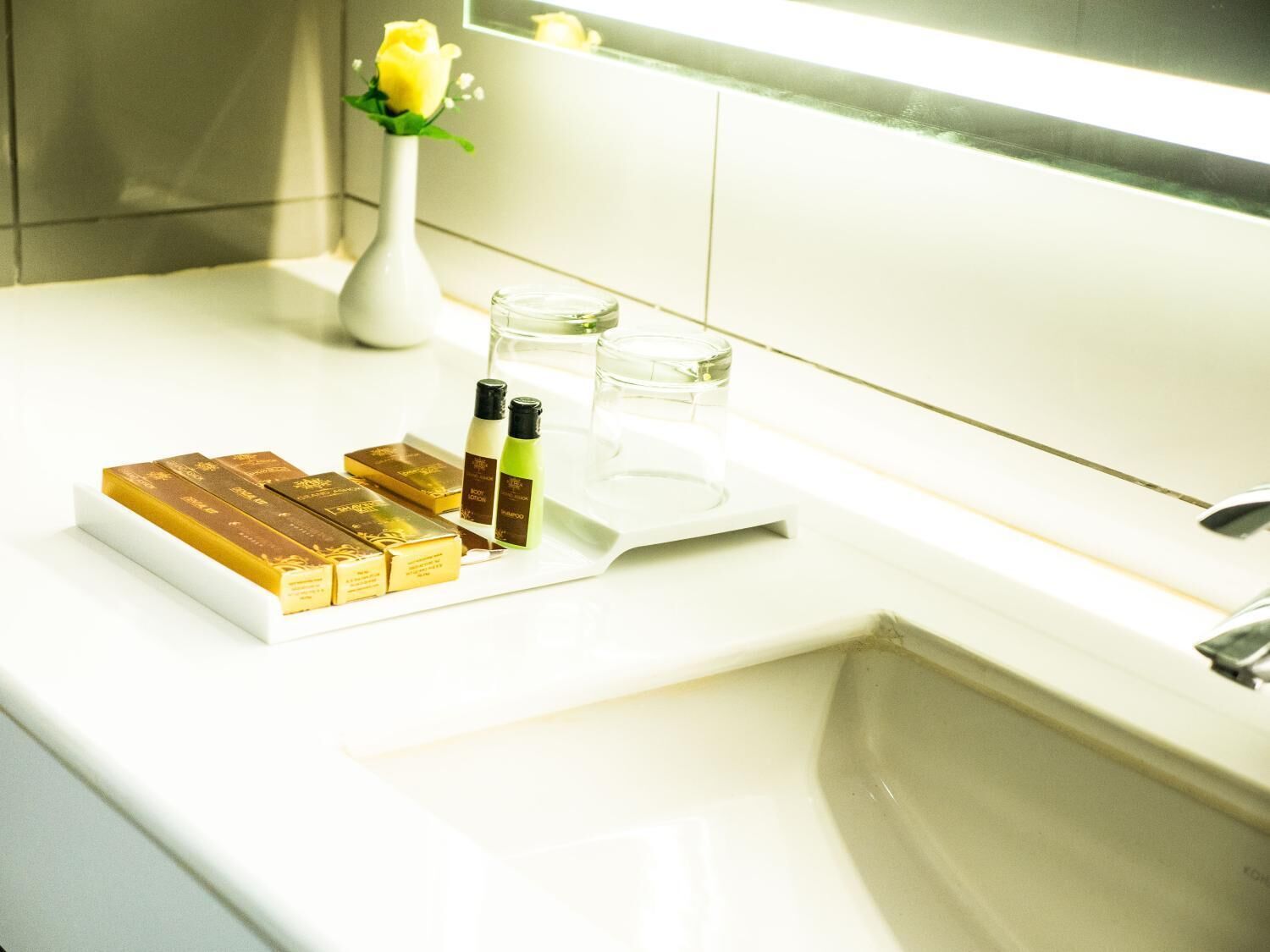 A bathroom counter with a white sink, a tray holding gold bars, two bottles of lotion, and a vase with a yellow rose.
