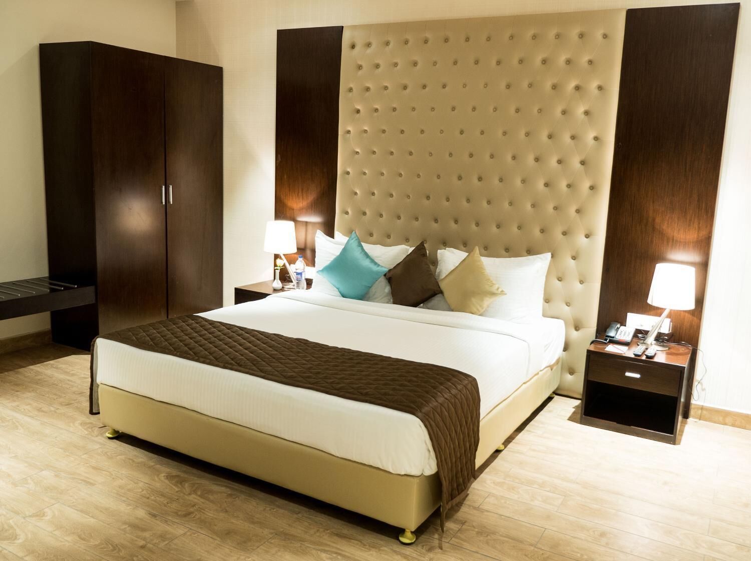 Una habitación de hotel con una cama grande, un armario, una mesita de noche con lámpara y otra mesita con teléfono y controles remotos.
