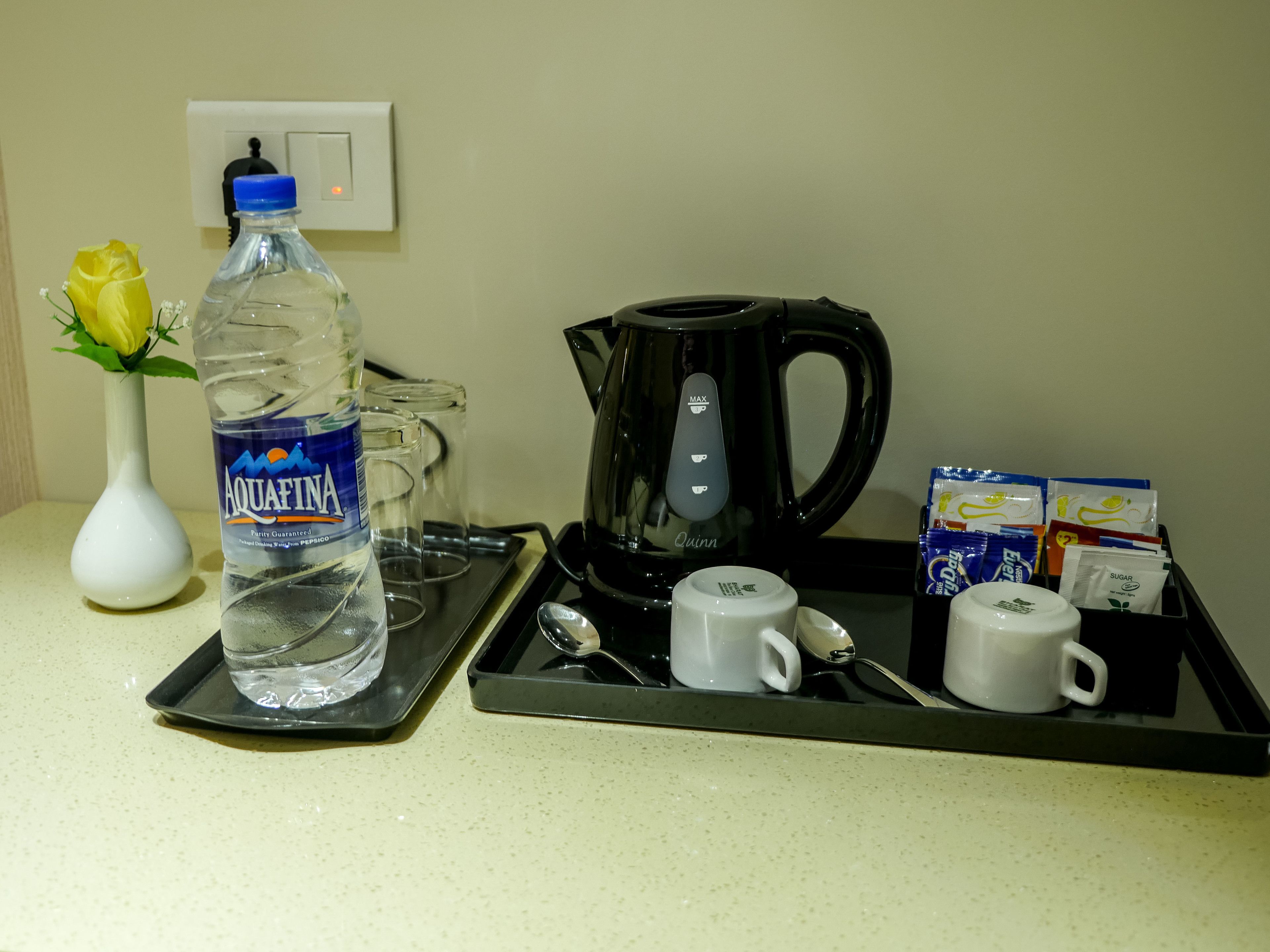 Ontbijtplateau in een hotelkamer met een fles Aquafina water, een witte mok, een zwarte koffiezetapparaat en suikerzakjes.