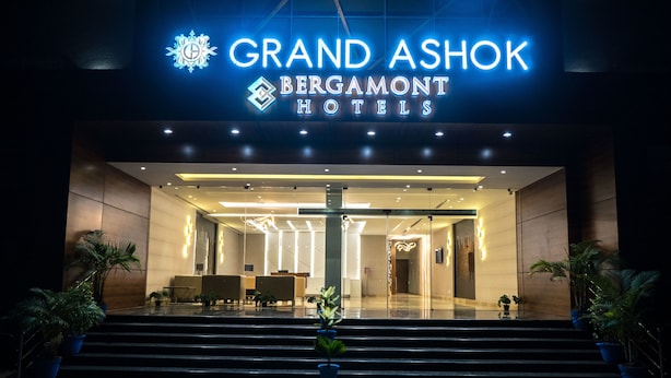 Entrada del Grand Ashok Bergamot Hotels, con un diseño moderno, que destaca un letrero iluminado, una zona de recepción y una escalera hacia el vestíbulo.