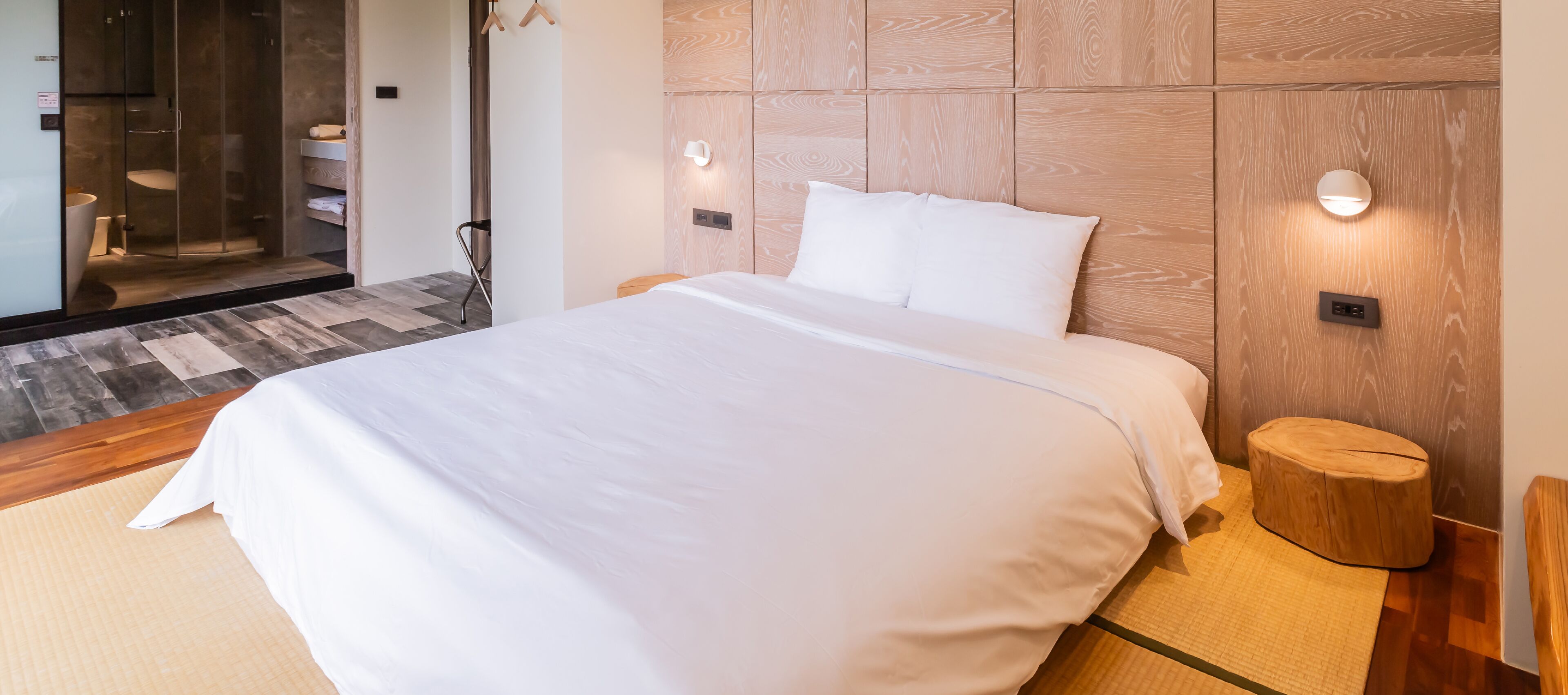 A modern hotel room with a large bed, a wooden headboard, bedside lamps, and a small wooden stool.