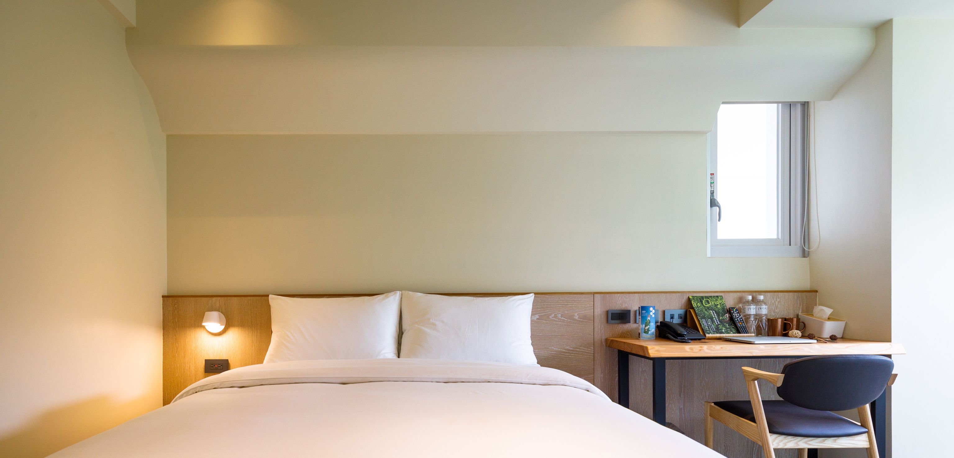 Ein Hotelzimmer mit Bett, Schreibtisch mit Stuhl, Fenster und Nachttisch mit Lampe.