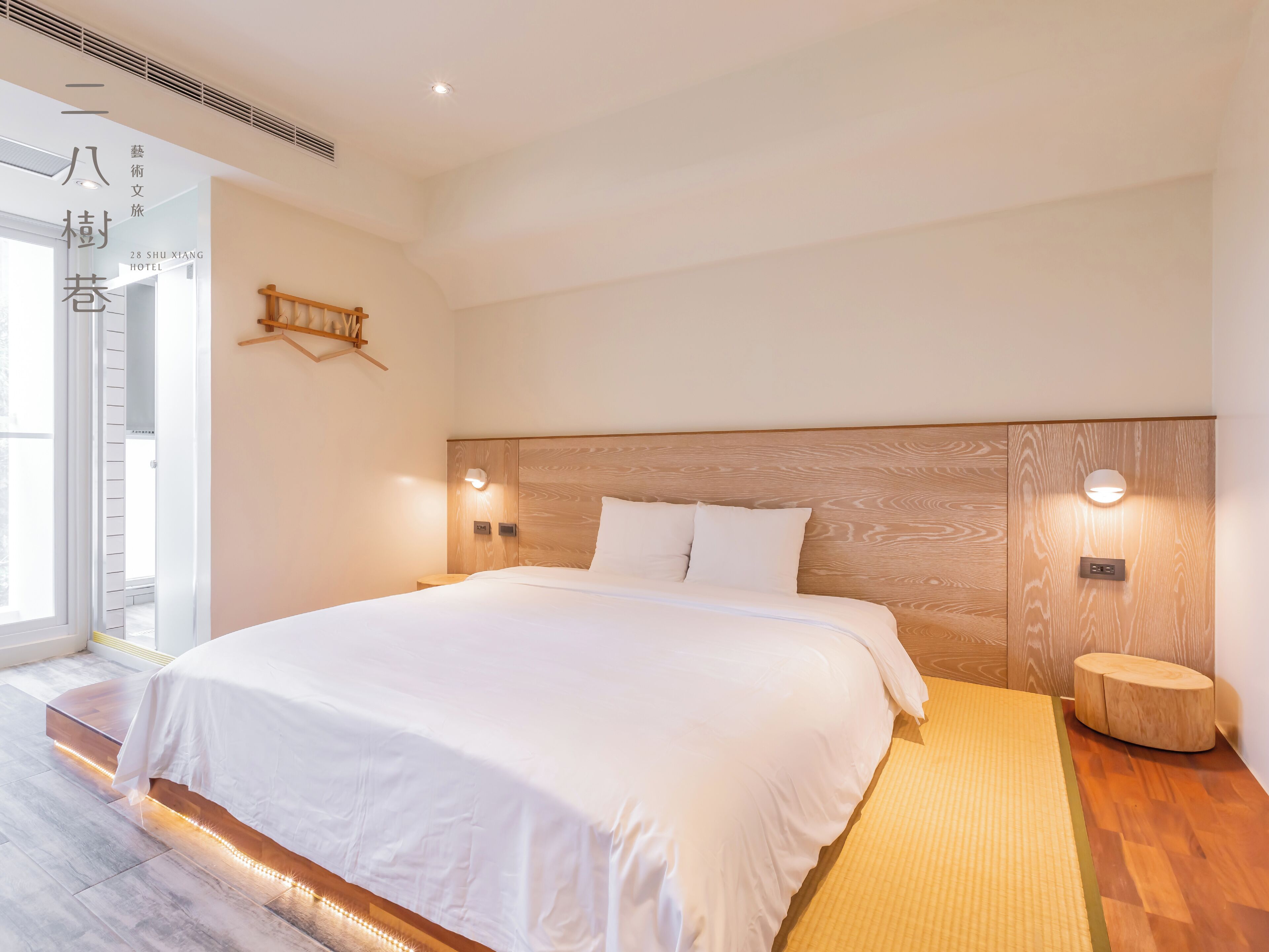 A modern hotel room with a large bed, wooden headboard, bedside lamps, and a small wooden stool.