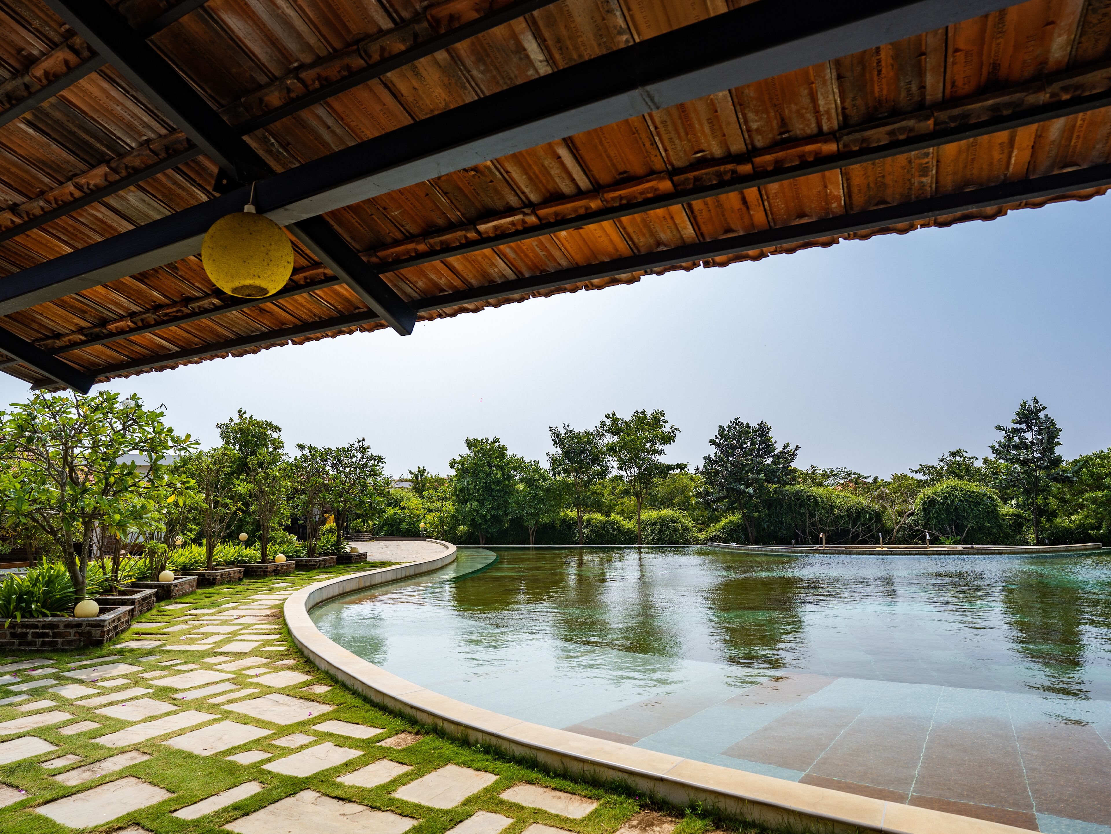 A covered outdoor area with a curved pool, stone pathway, and lush greenery.