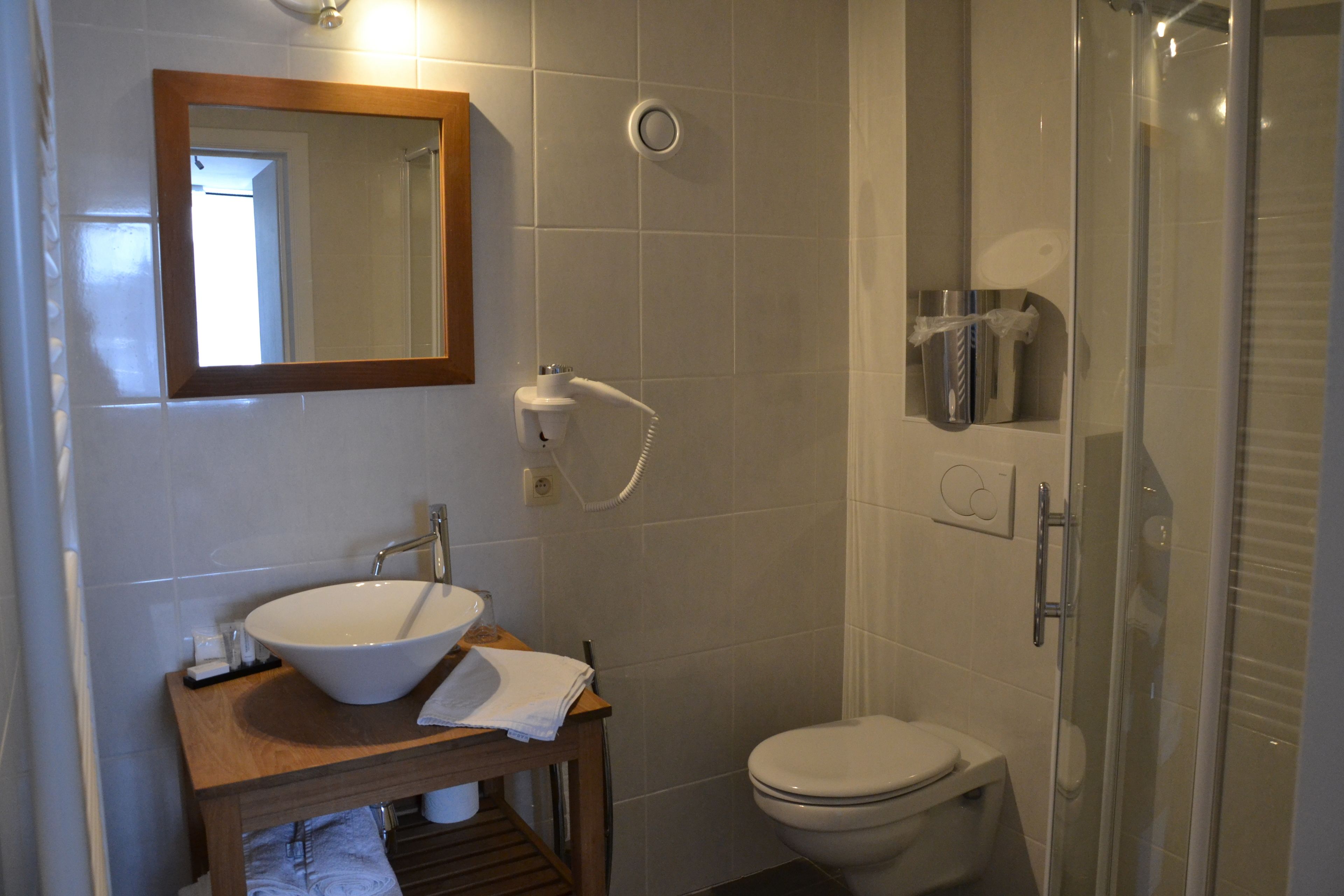 Un bany amb un tocador de fusta, un lavabo blanc, un mirall, un vàter i una zona de dutxa amb envàs de vidre.