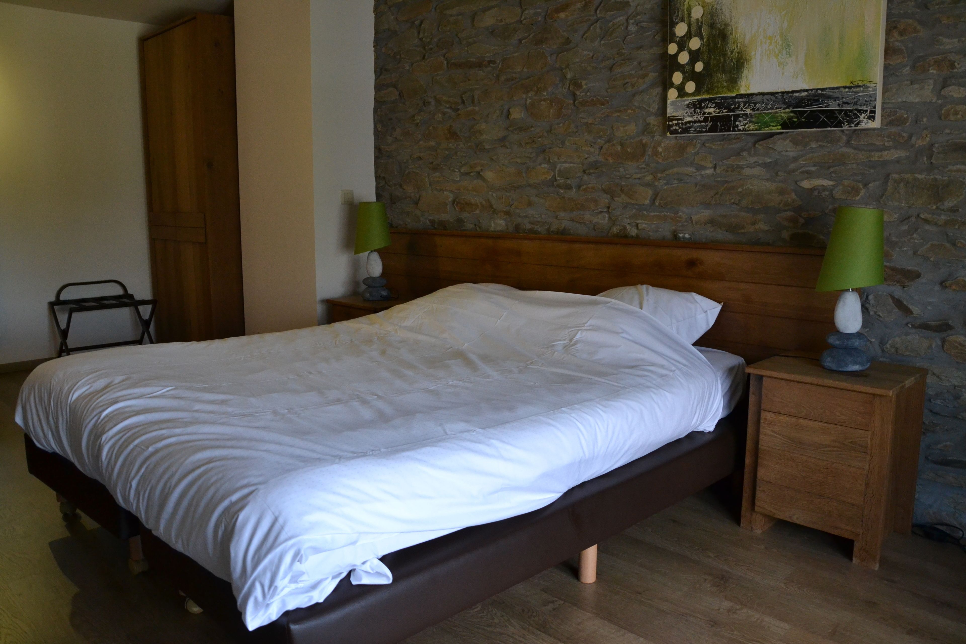 A bedroom with a wooden bed, a nightstand with a lamp, a wardrobe, and a stone wall.