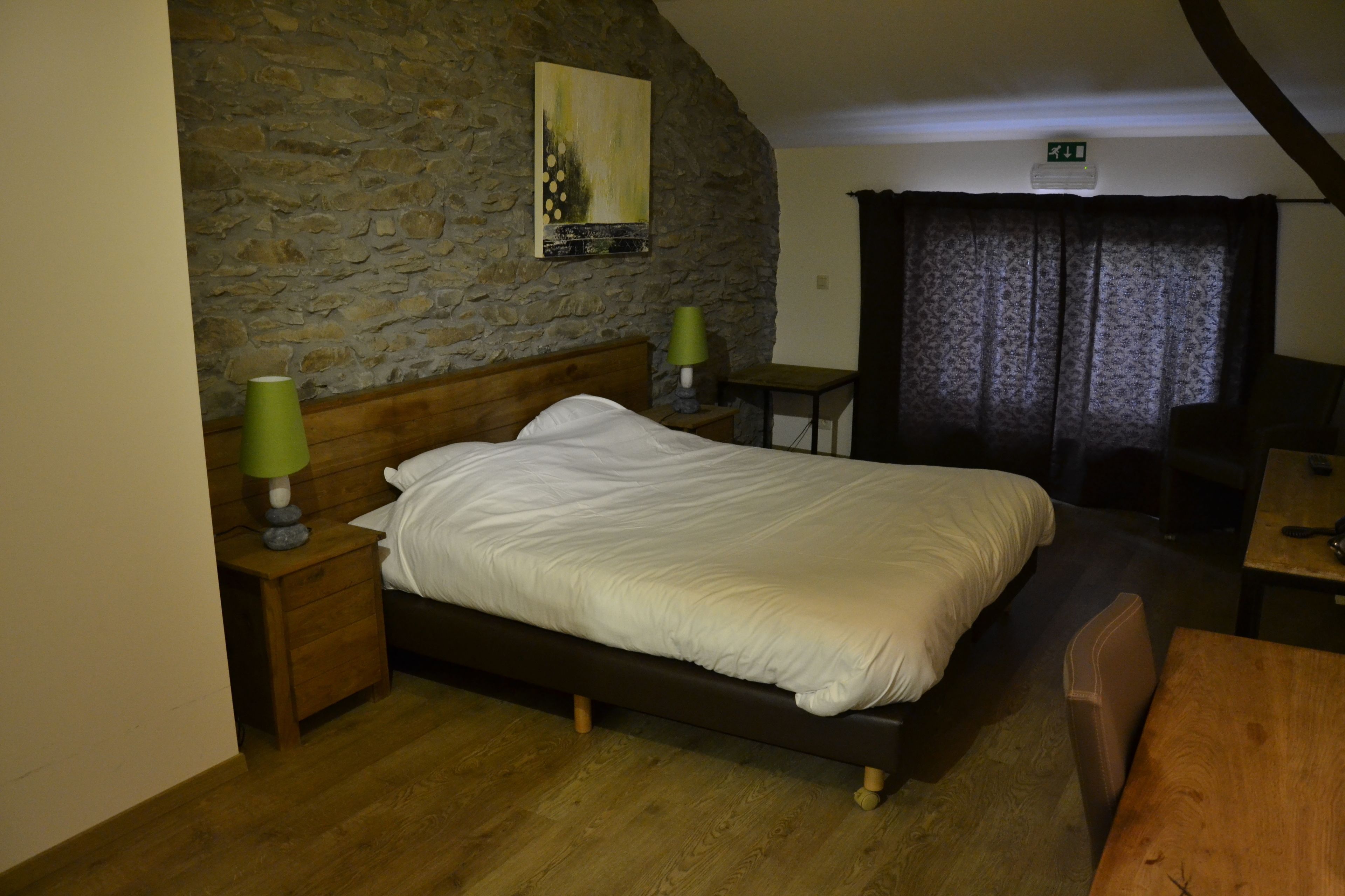 Une chambre avec un mur en pierre, un lit avec une literie blanche, une table de chevet en bois avec une lampe, et une petite table avec une lampe.