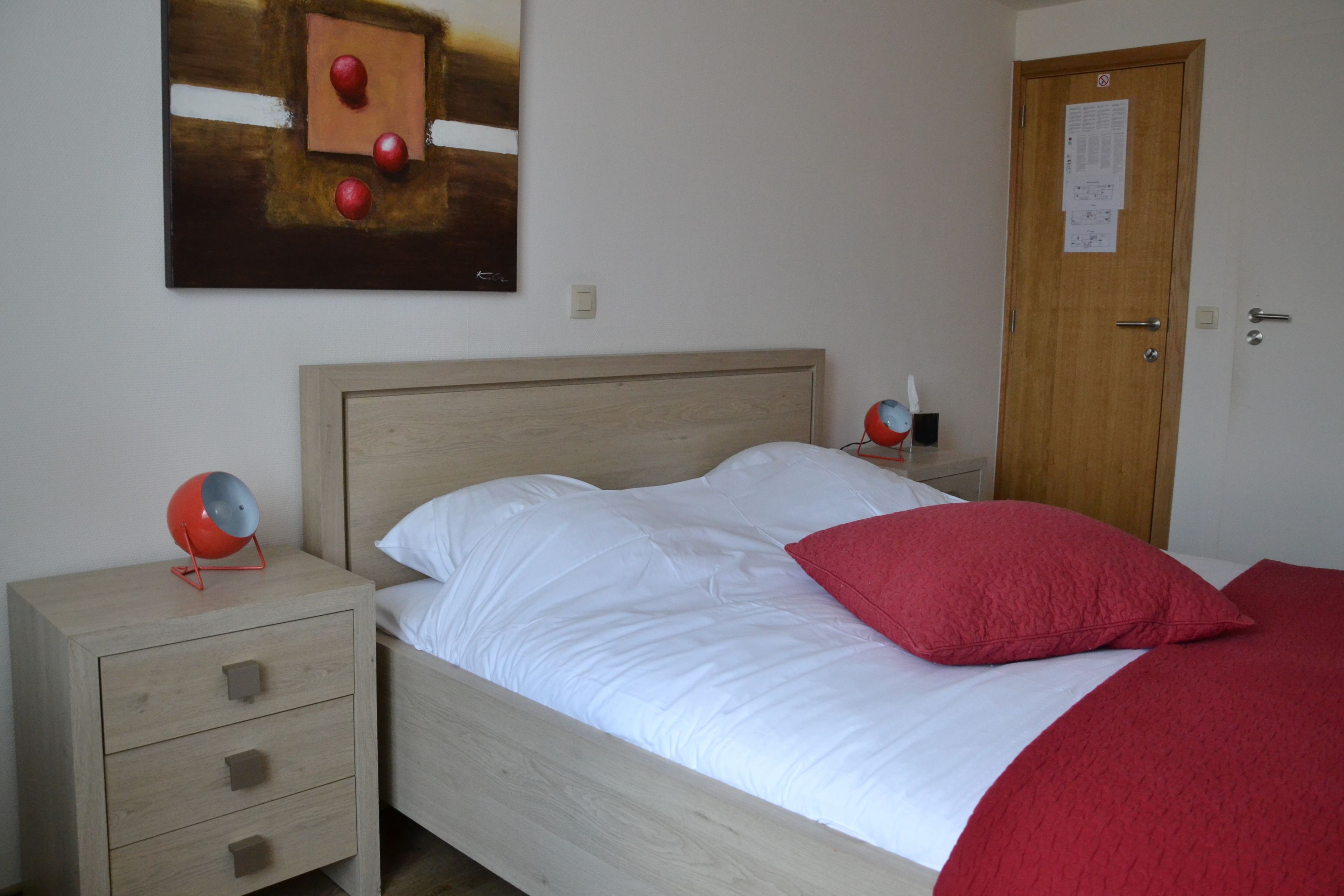 Une chambre à coucher avec un lit en bois, une table de chevet avec des tiroirs, une lampe rouge et un tableau accroché au mur.