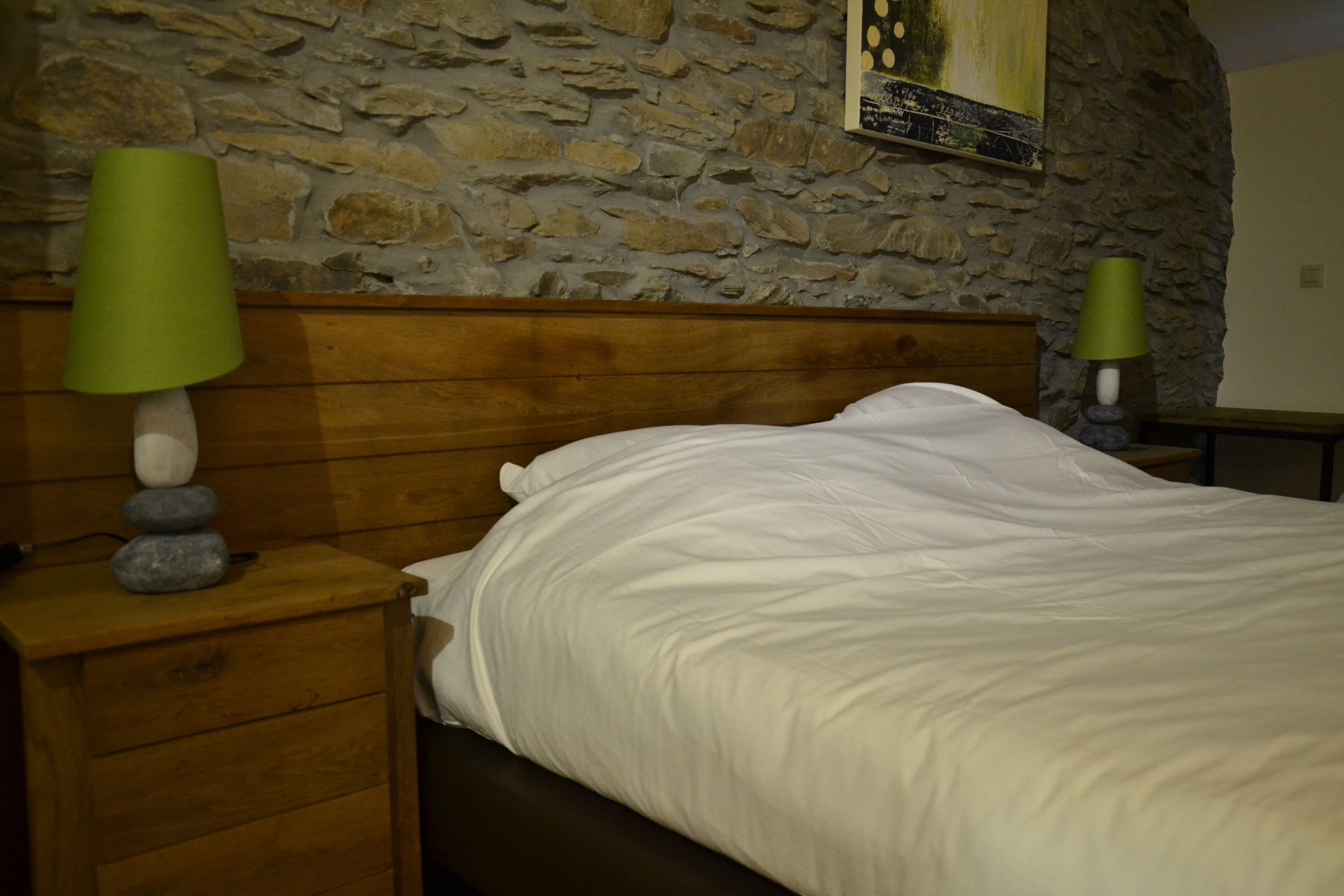 A bedroom with a stone wall, wooden headboard, a bed with white bedding, and two green lamps on wooden nightstands.