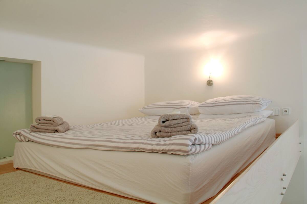 A neatly made bed with striped bedding and two pillows, a wall-mounted light fixture, and a door to the left.