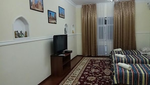 A hotel room with a TV on a wooden stand, a patterned rug, a bed with striped bedding, and a window with curtains. - Hotel Abu Shakheen (Bukhara)
