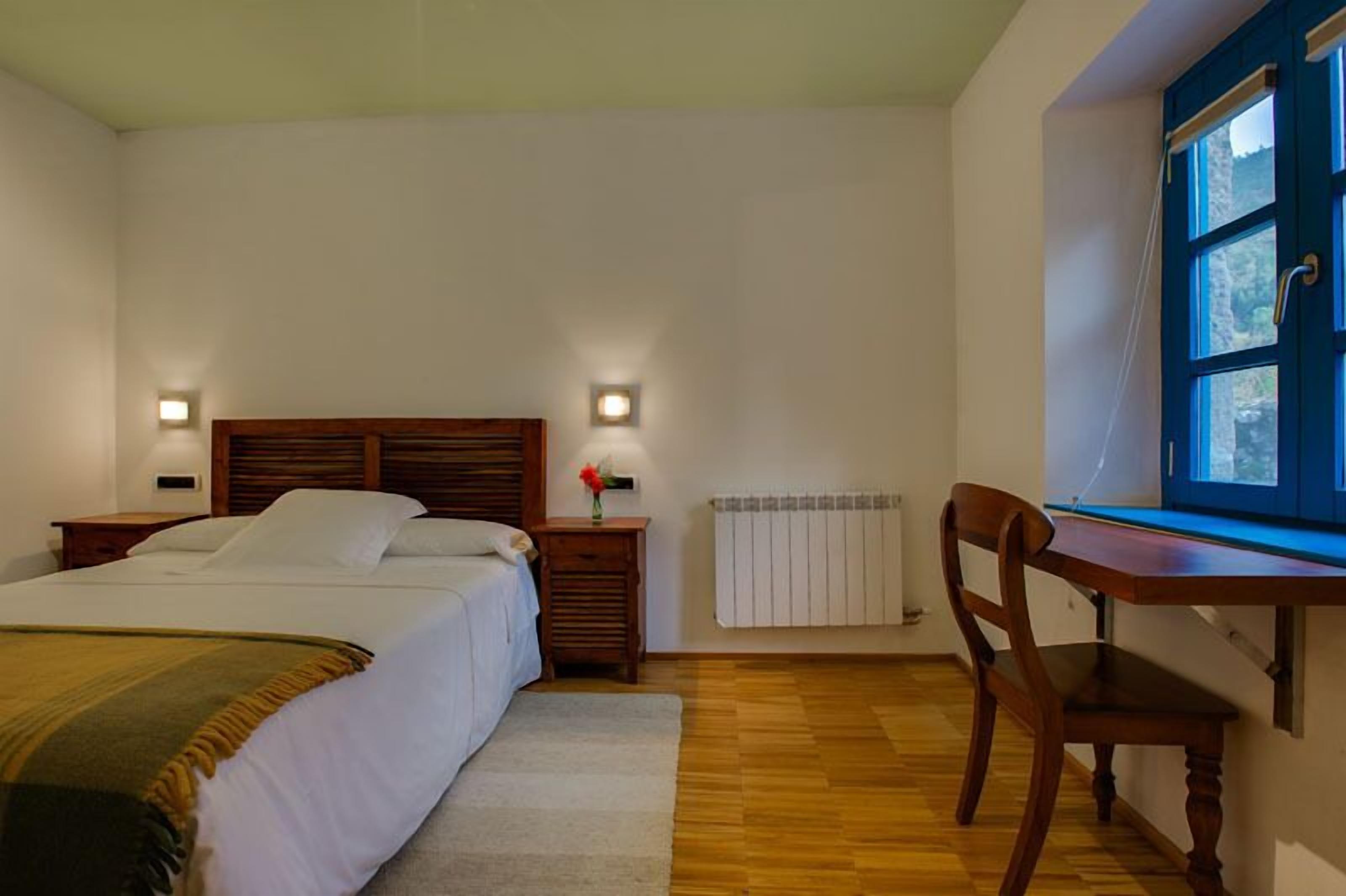 Un dormitorio con cama, mesitas de noche, una silla, ventana y radiador.