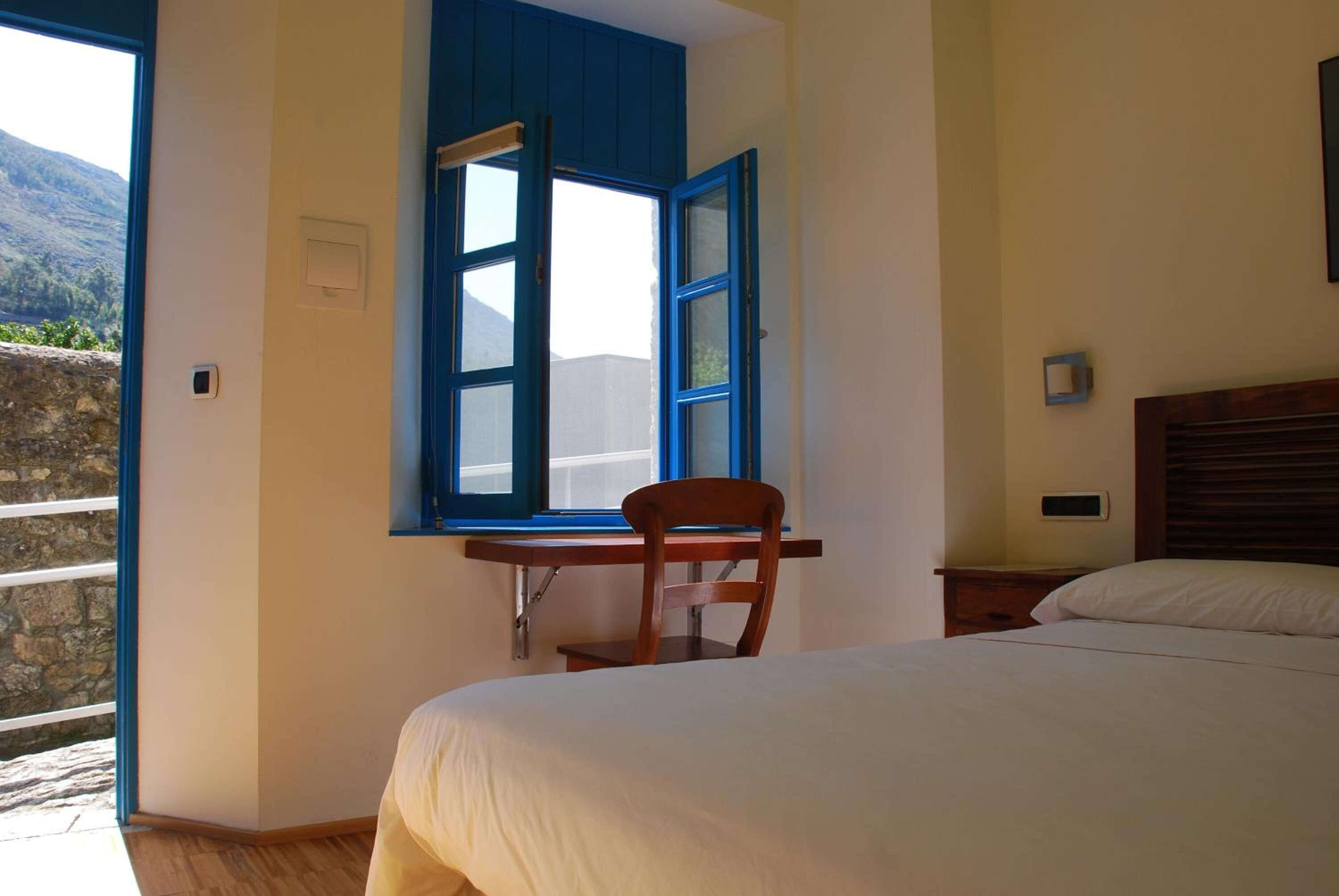 Un dormitorio con una cama, una silla de madera, un escritorio con silla, una ventana con contraventanas azules y vistas a un paisaje montañoso.