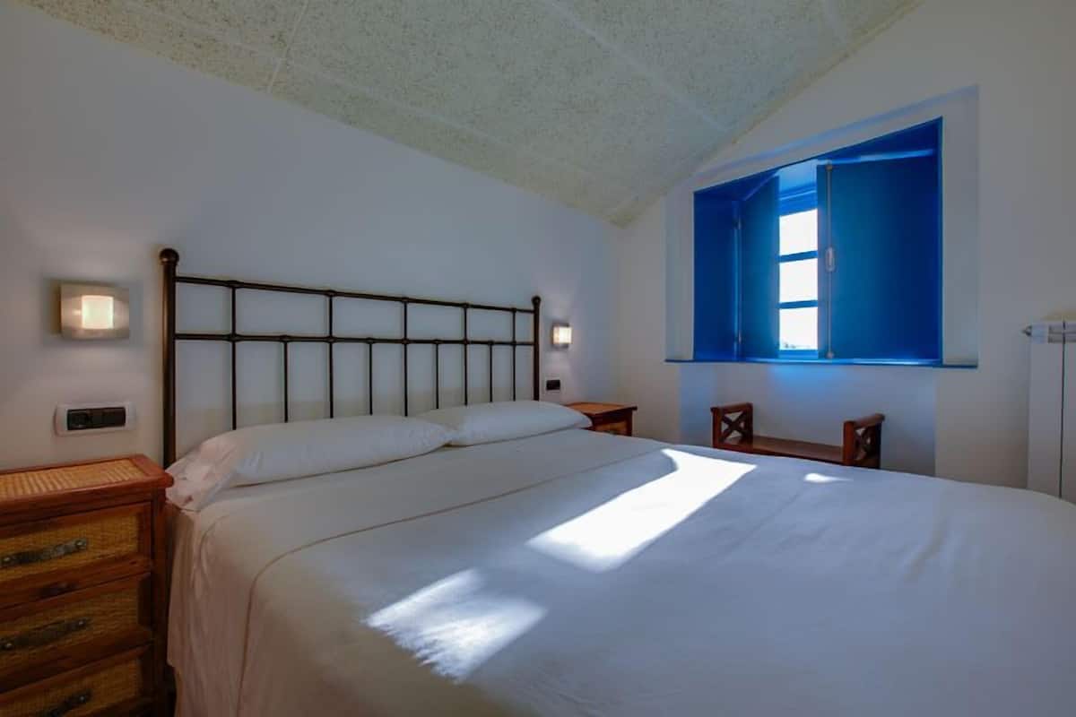 Un dormitorio con una cama grande, cabecera de madera, lámparas de noche, mesita de noche y una ventana con contraventanas azules.
