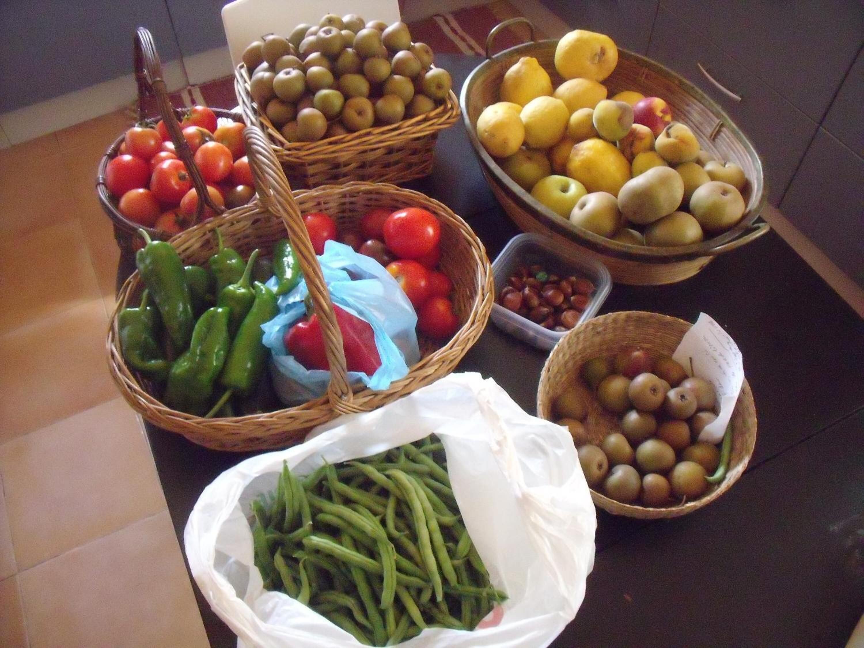 A variety of fresh produce including tomatoes, green peppers, apples, lemons, and green beans displayed in baskets and a plastic bag.