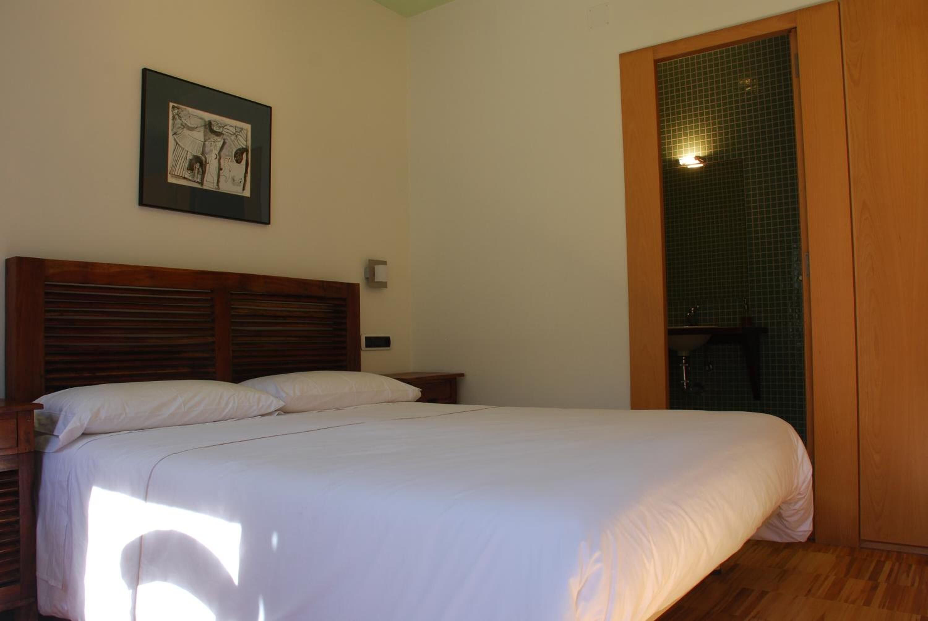 A bedroom with a wooden headboard, a bed with white linens, a framed picture on the wall, and a green-tiled shower area.