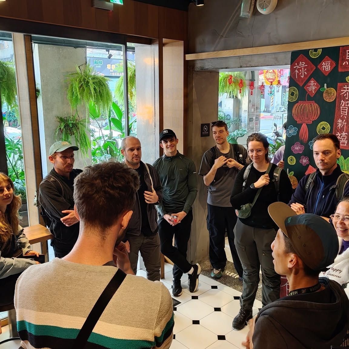 A group of people in a cafe with a large window, indoor plants, and a wall with Chinese characters.