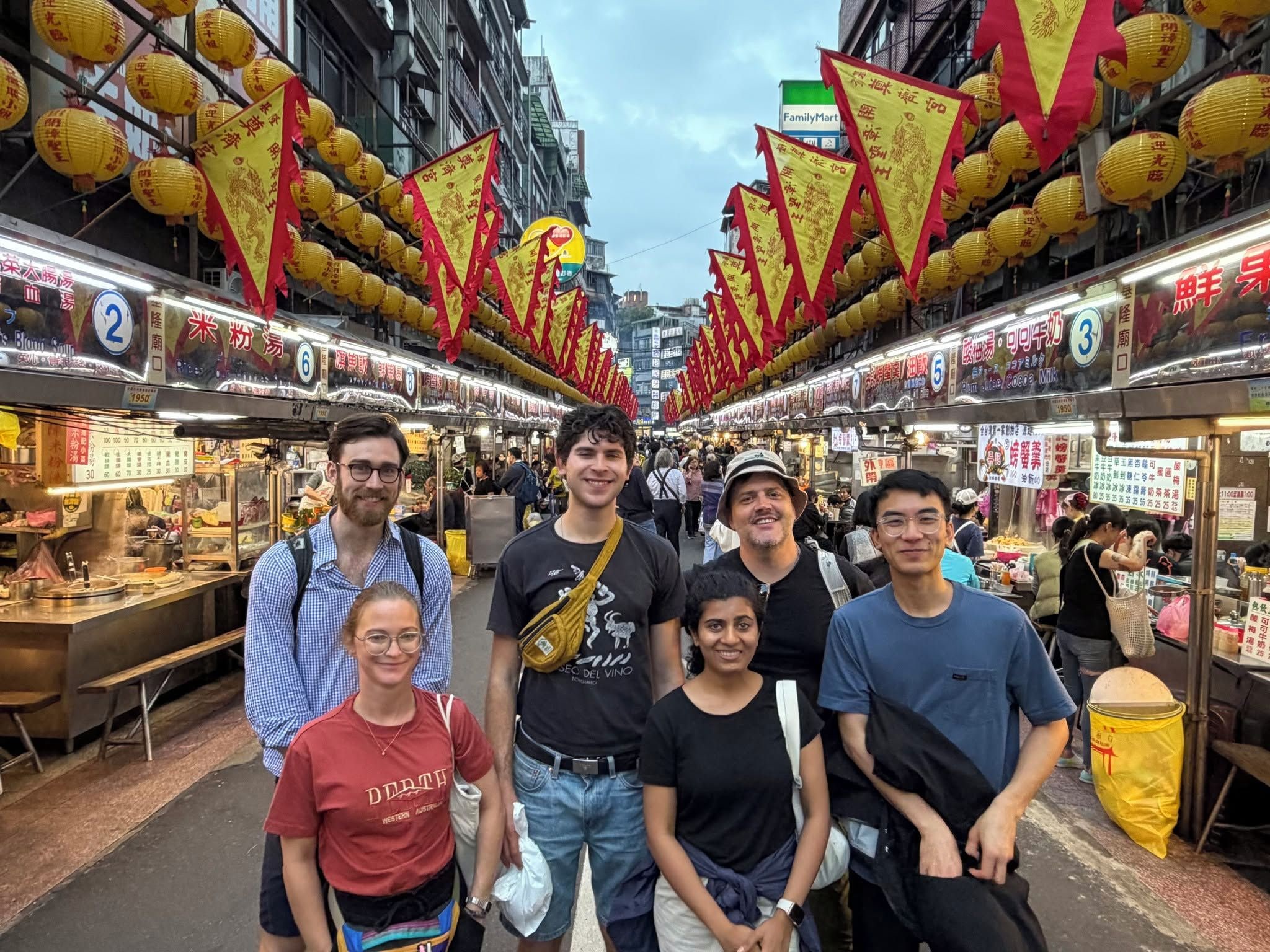 Een groep mensen staat op een markt met kraampjes aan weerszijden, onder een overkapping van gele lampionnen.