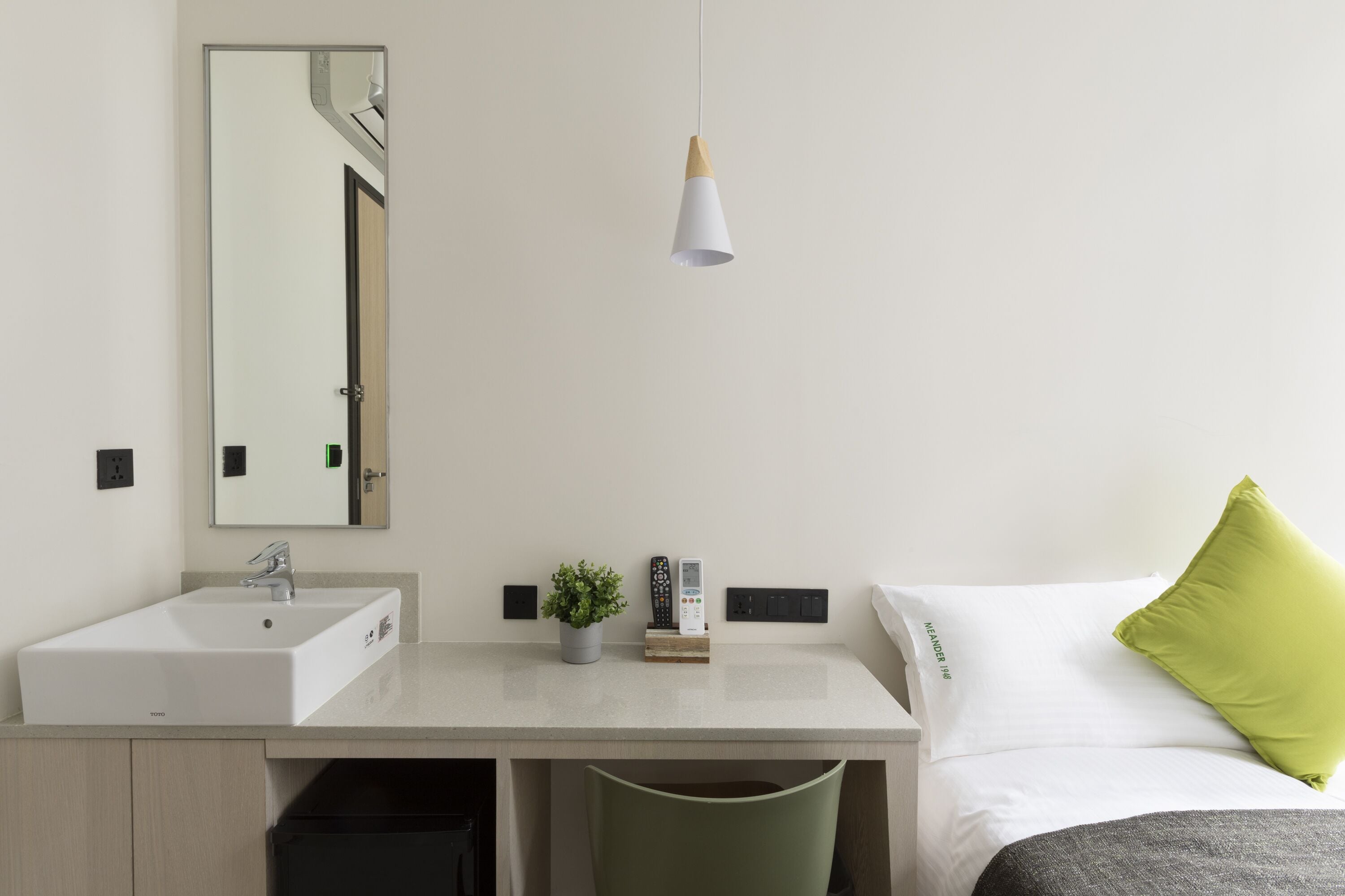 A hotel room with a bed, a bedside table with a lamp, a mirror, and a small potted plant.