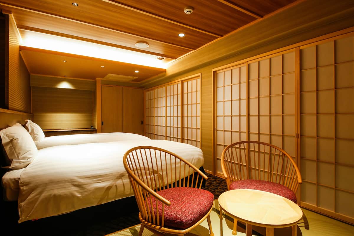 Une chambre d’hôtel avec un grand lit, deux chaises en bois, une petite table et des paravents coulissants de style japonais.