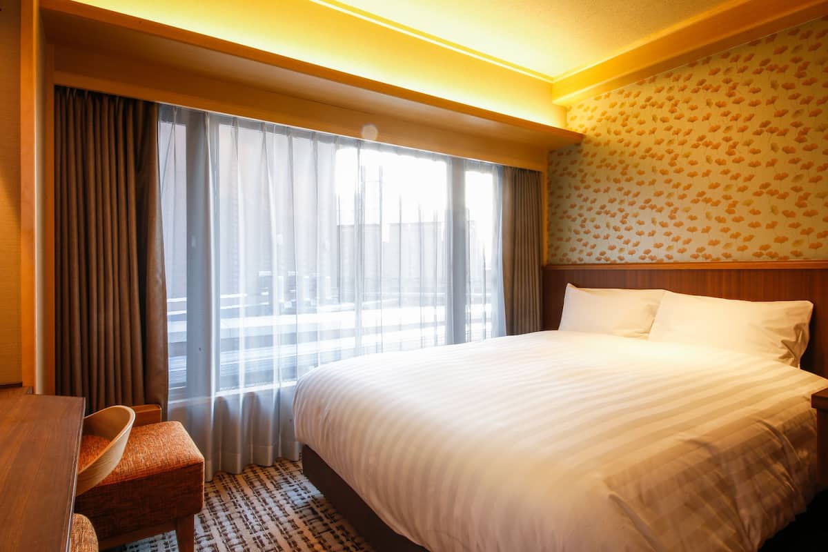 Une chambre d’hôtel avec un grand lit, une tête de lit en bois, une table à manger avec une chaise et une fenêtre avec des rideaux.