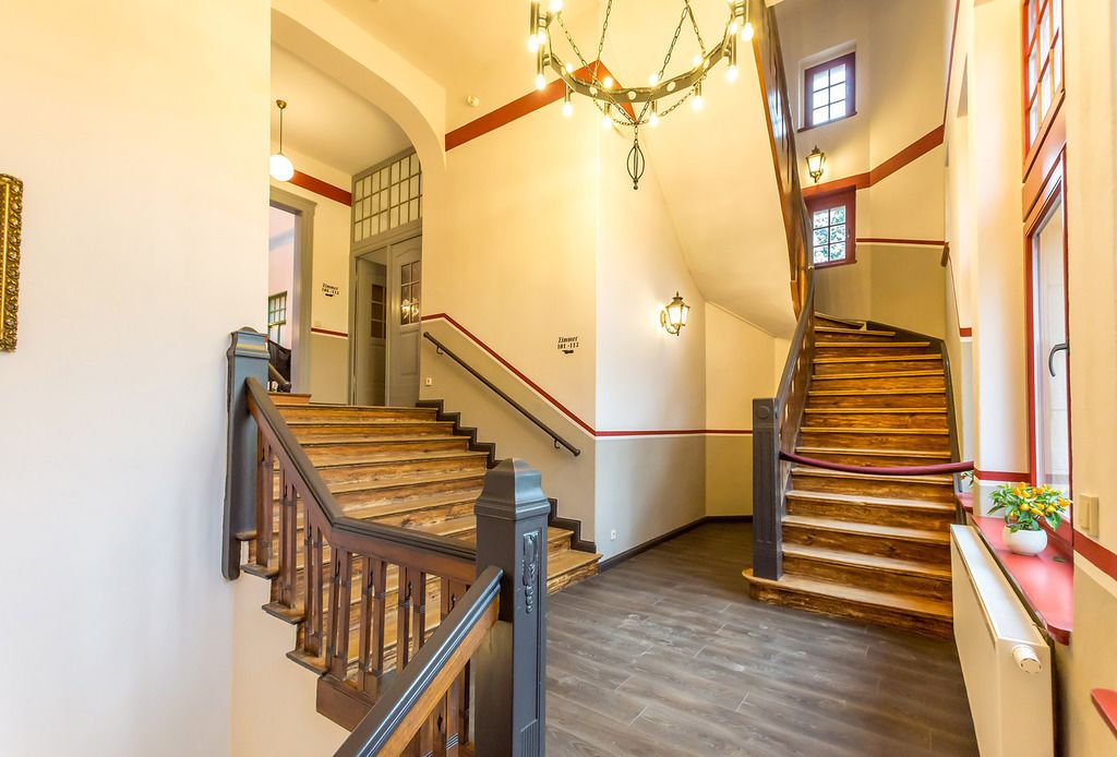 A grand staircase with wooden steps and a red railing, leading to a landing with a chandelier.