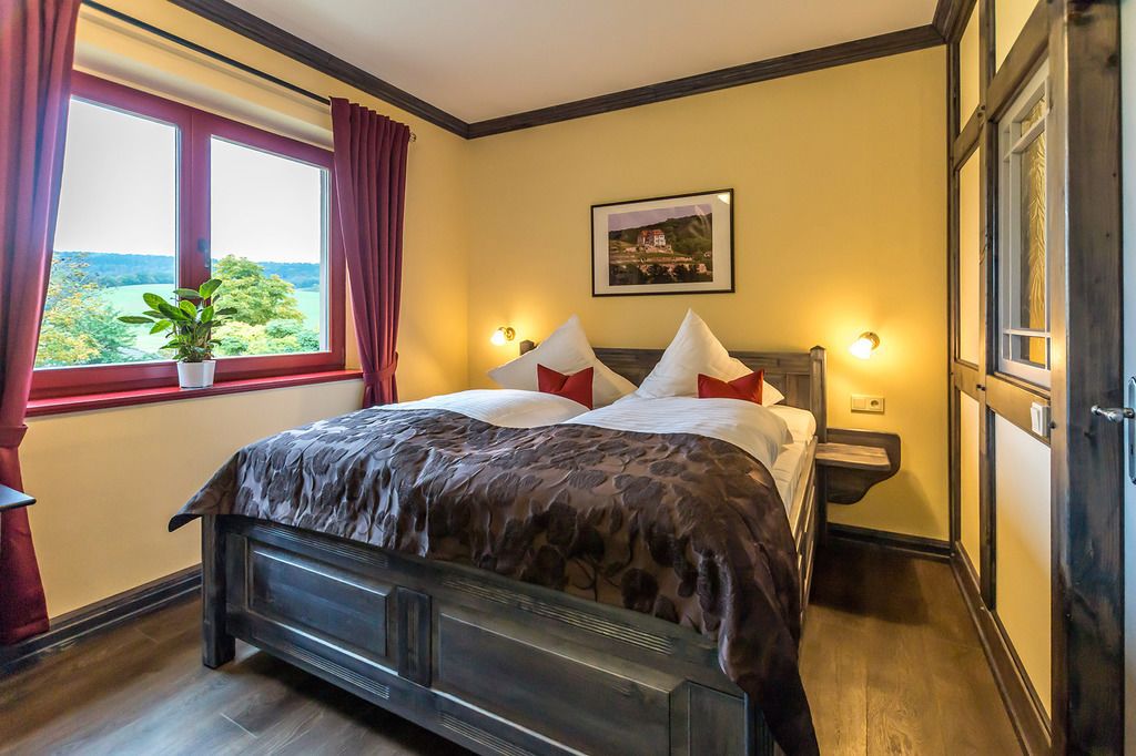 Ein Schlafzimmer mit einem großen Bett, einem Fenster mit Vorhängen, einem Nachttisch mit Lampe und einem gerahmten Bild an der Wand.