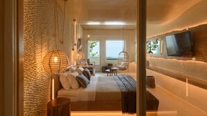 A modern bedroom with a large bed, a sofa, a TV mounted on the wall, and a view of the outdoors. - Divelia East (Santorini)
