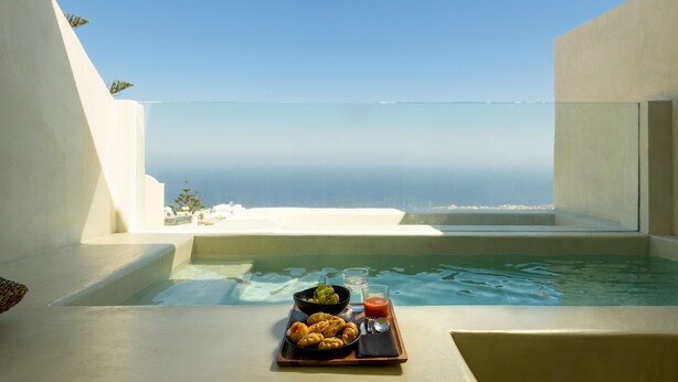 A balcony with a view of the sea, a hot tub, and a tray with food and drinks. - Divelia East (Santorini)