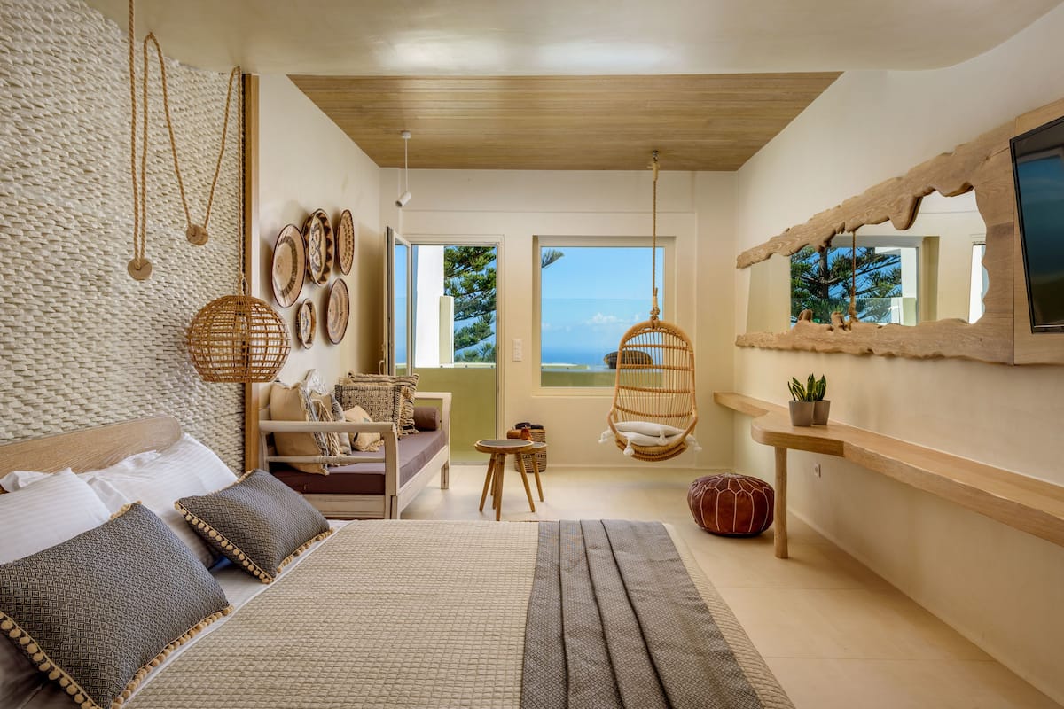A modern bedroom with a large bed, a hanging chair, a small table, and a view of the ocean.