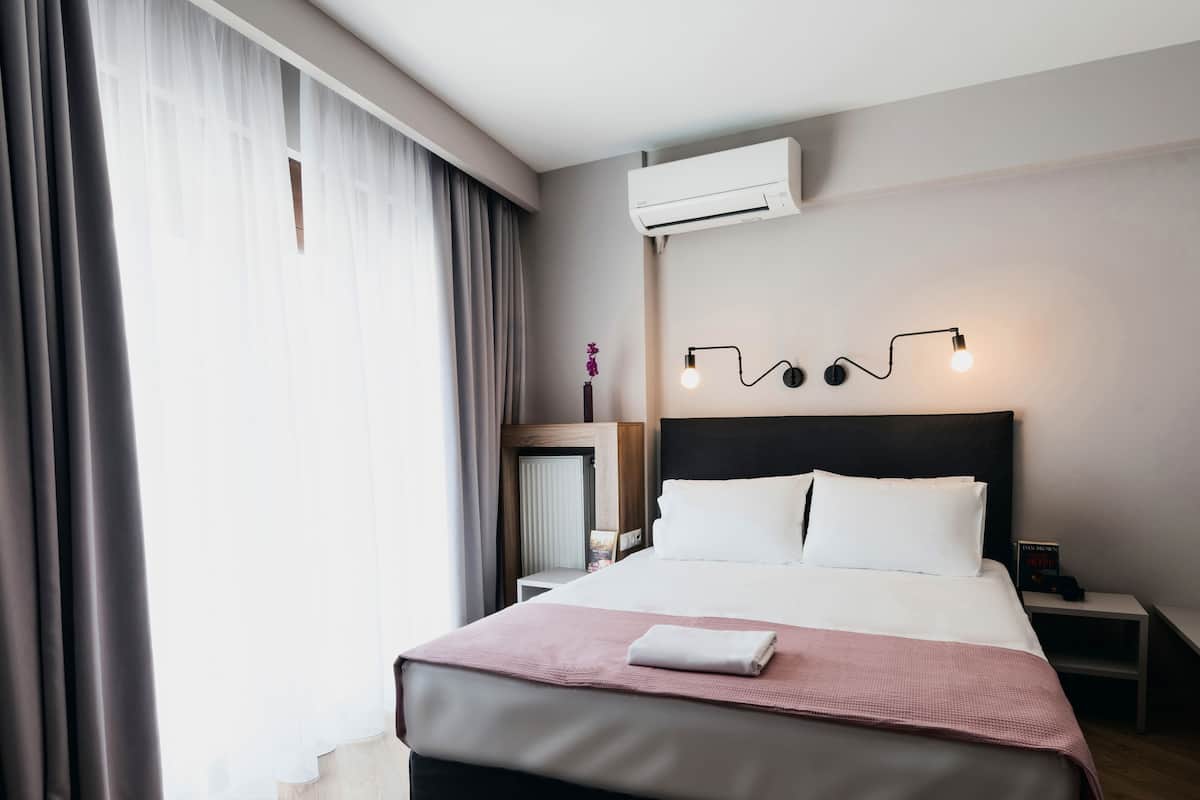 Una habitación de hotel moderna con una cama grande, una mesita de noche, un aire acondicionado y una ventana con cortinas.