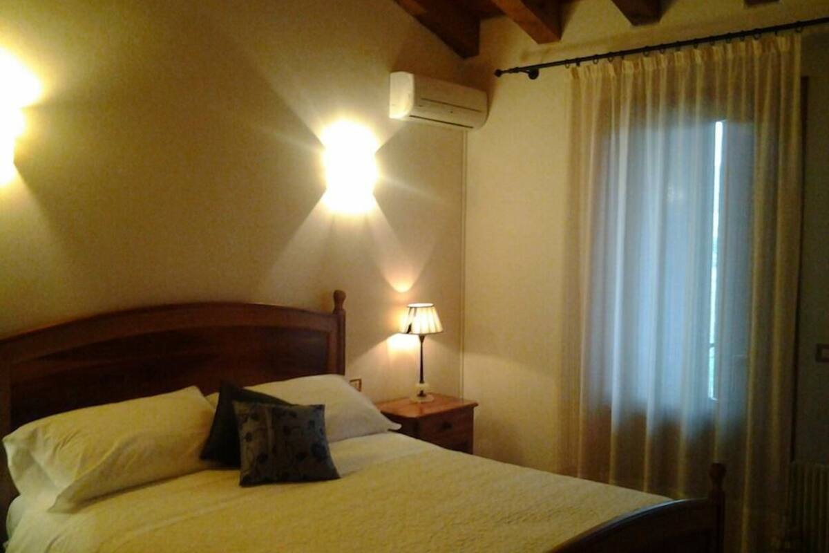 Un dormitorio con una cama de madera, una mesita de noche con una lámpara, un aire acondicionado instalado en la pared y una ventana con cortinas.