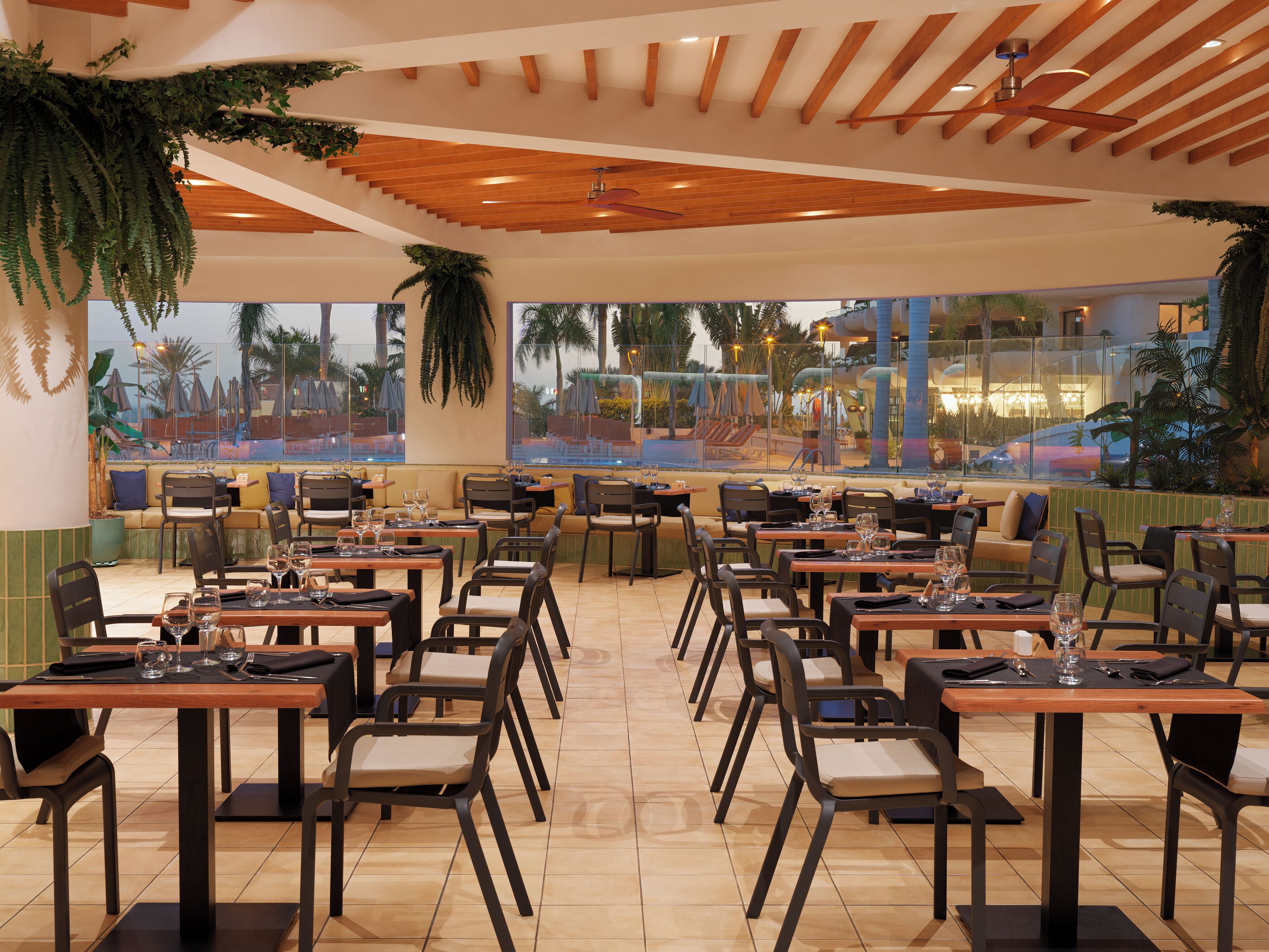 Un restaurante con varias mesas y sillas, amplias ventanas que ofrecen vistas a una piscina y palmeras.