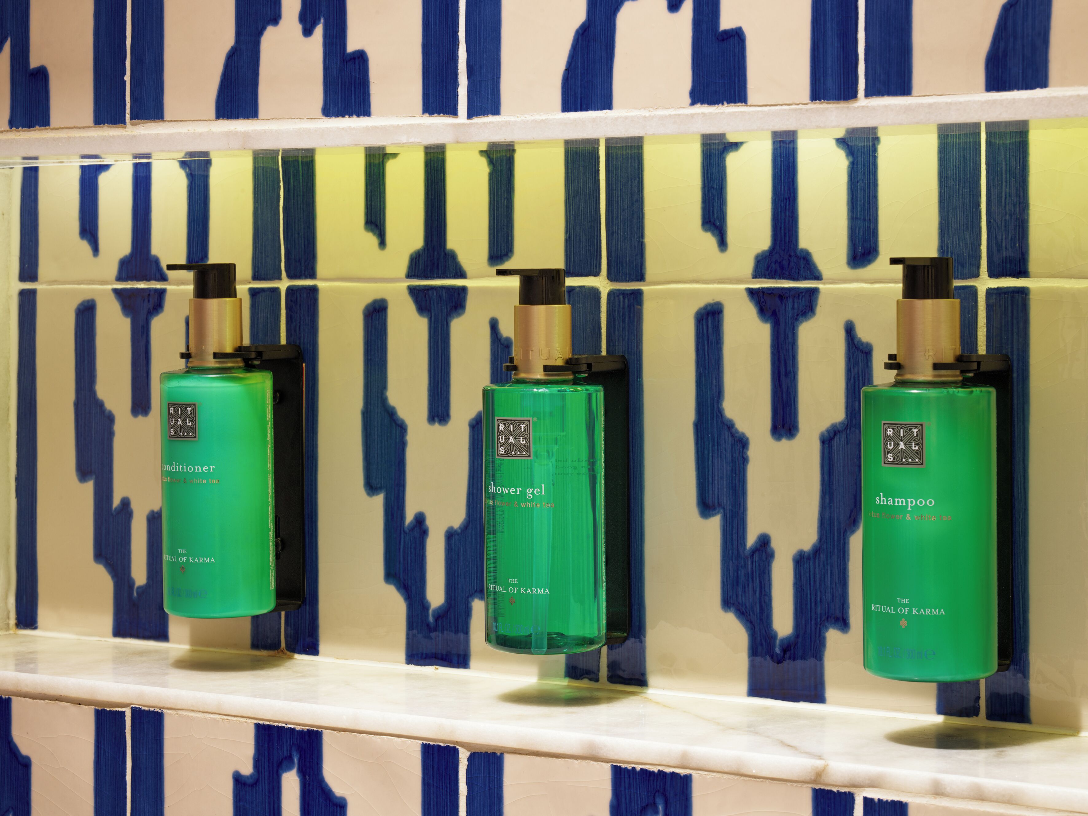 Tres botellas verdes de productos 'Ritual of Karma' sobre una estantería frente a una pared con azulejos azules y beige.