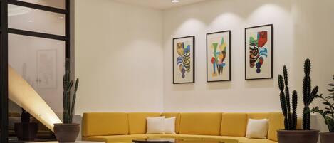 A modern lobby with a yellow sofa, a coffee table, and three framed artworks on the wall.