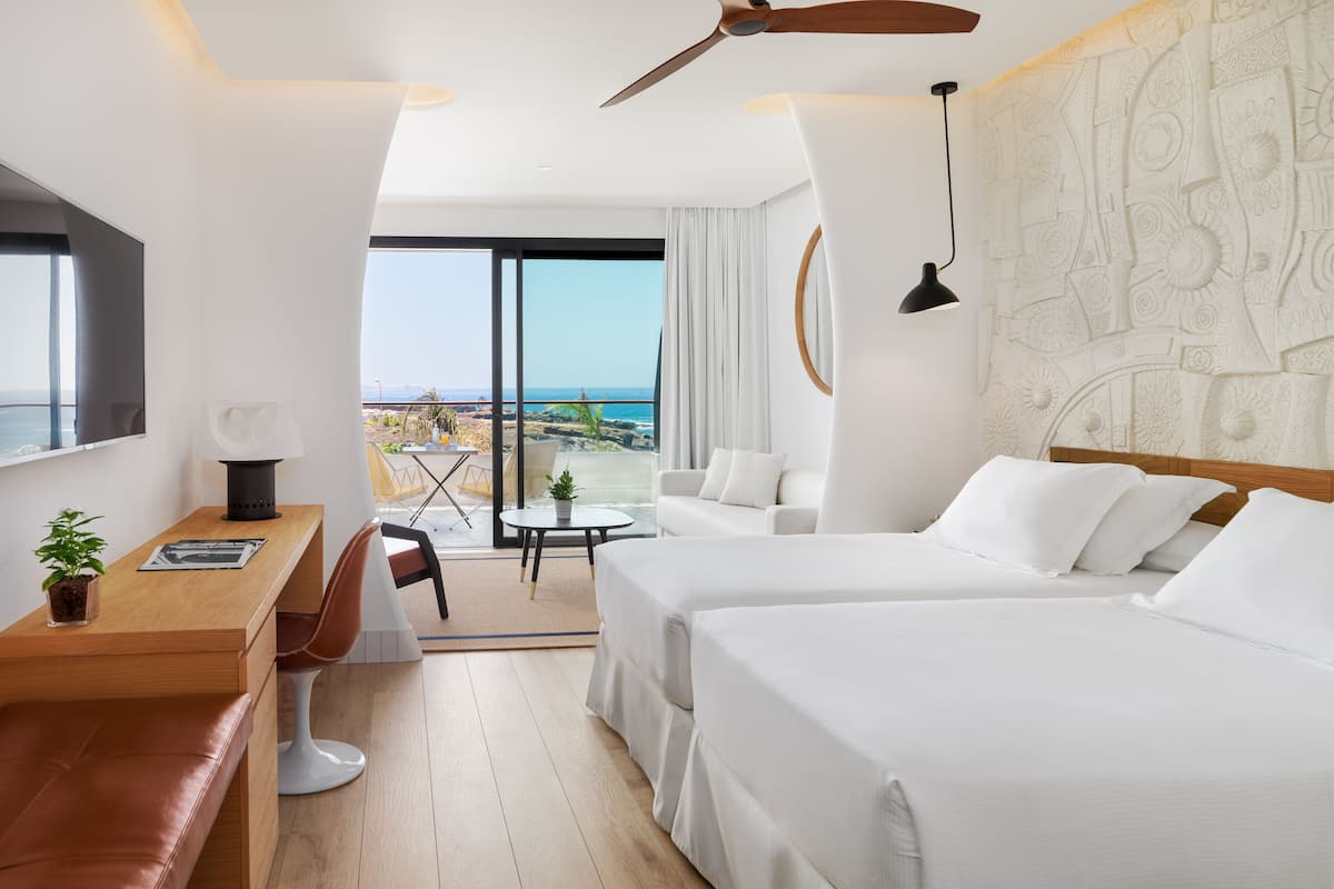 A hotel room with two beds, a desk, a chair, a small table, and a balcony with a view of the ocean.