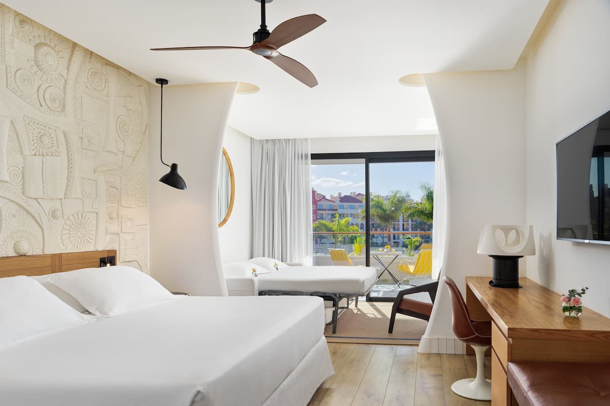 A hotel room with a bed, a desk, a chair, a ceiling fan, a TV, and a balcony with a view of buildings and palm trees.